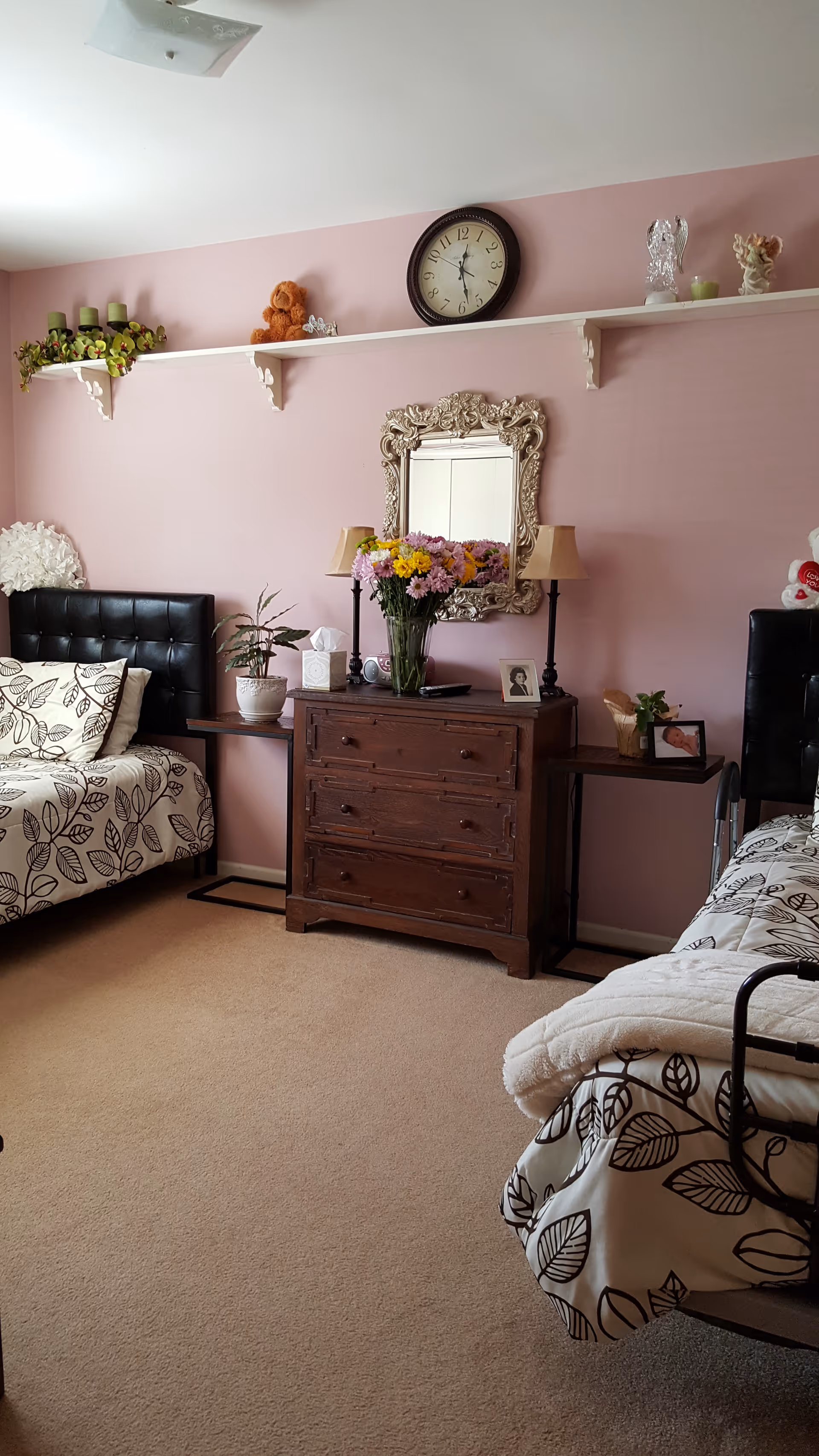 A cozy bedroom with two single beds featuring black tufted headboards and leaf-patterned bedding. Between the beds is a wooden dresser with a vase of colorful flowers, two lamps, and framed photos. Above the dresser is an ornate mirror and a wall clock. A white shelf runs along the wall holding decorative items including candles, a teddy bear, and small statues. The walls are painted pink and the floor is carpeted.