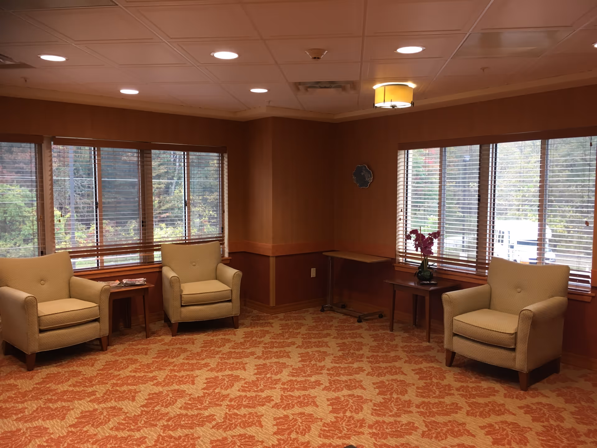 A cozy sitting area with three beige armchairs arranged around two small wooden side tables, one of which has a vase with purple flowers. The room has large windows with wooden blinds letting in natural light, patterned carpet in warm tones, and a ceiling with recessed lighting and a hanging light fixture.