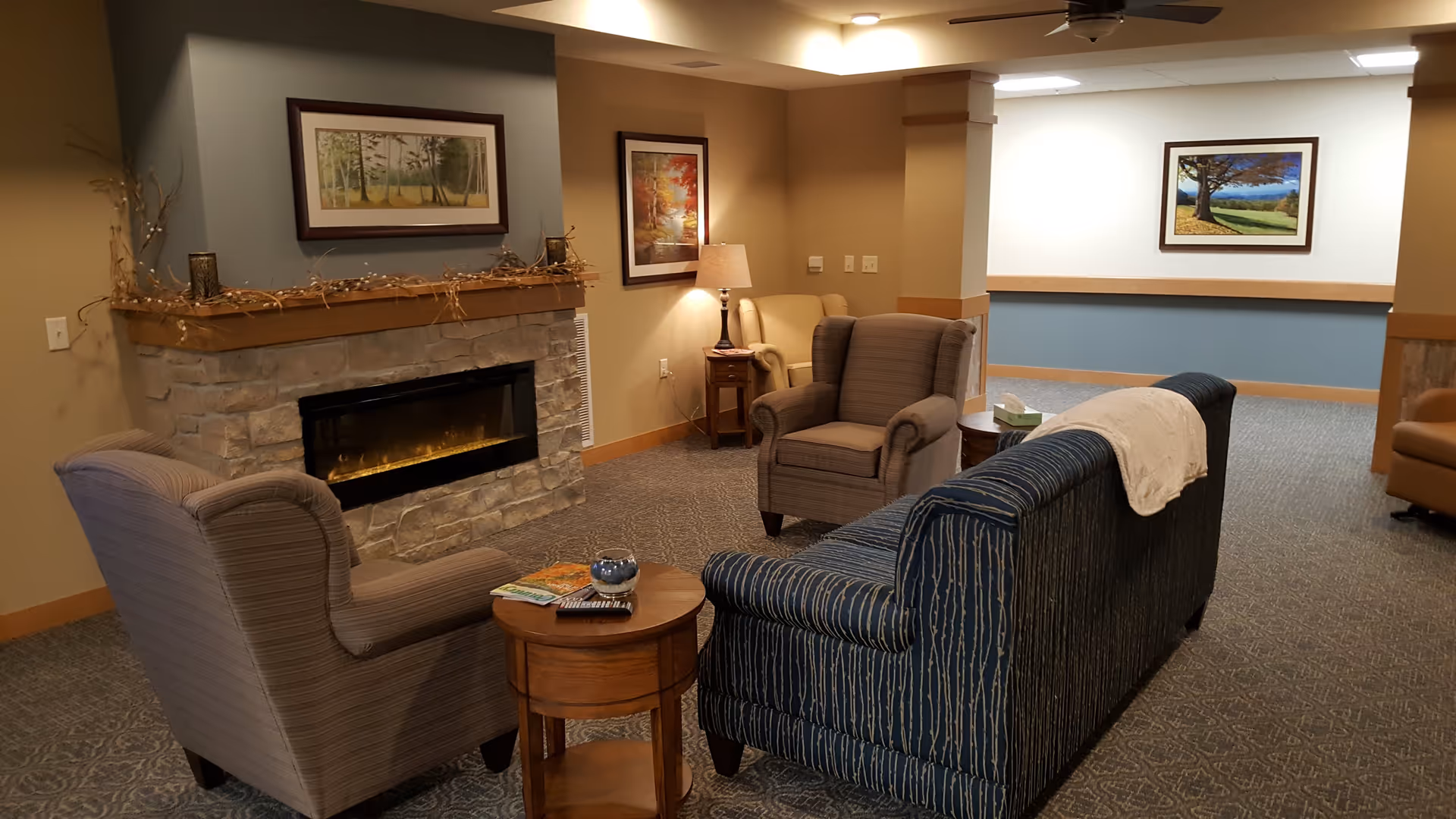 A cozy living room area in Oak Park Place Nakoma featuring a stone fireplace with a wooden mantel decorated with a garland. The room has a blue patterned sofa with a white throw draped over the back, two upholstered armchairs, a round wooden side table with magazines and a decorative item, and framed landscape paintings on the walls. The carpet is patterned and the lighting is warm and inviting.