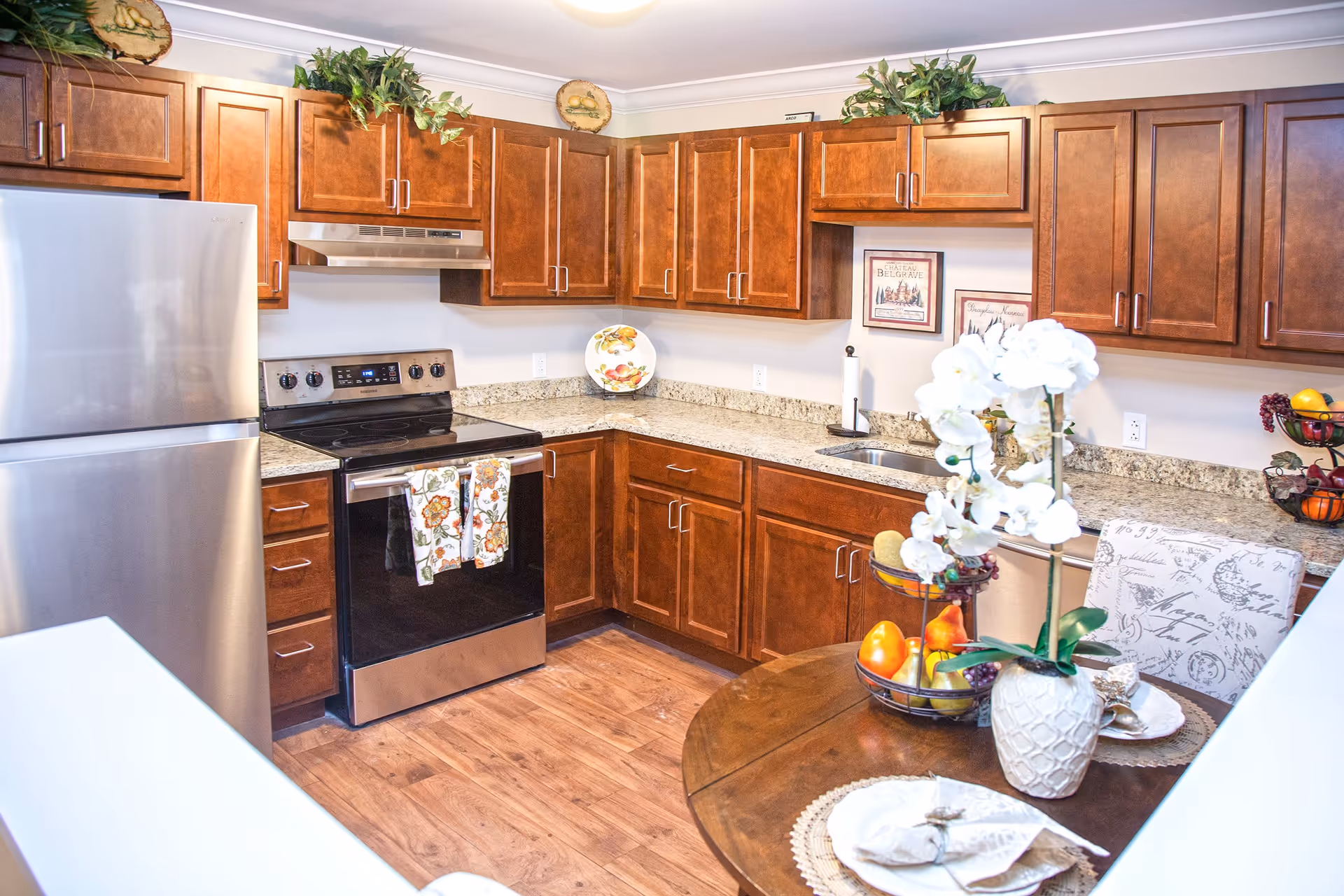 A bright kitchen with wooden cabinets, granite countertops, a stainless steel refrigerator, and an electric stove with two floral towels hanging on the handle. There is a round wooden table with a white orchid centerpiece, a fruit basket, and place settings. Decorative plates and framed pictures adorn the walls and countertop.