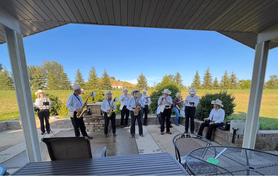 A group of musicians wearing white shirts and cowboy hats performing outdoors on a patio area with green grass and trees in the background. The scene is viewed from under a covered porch with patio furniture in the foreground.