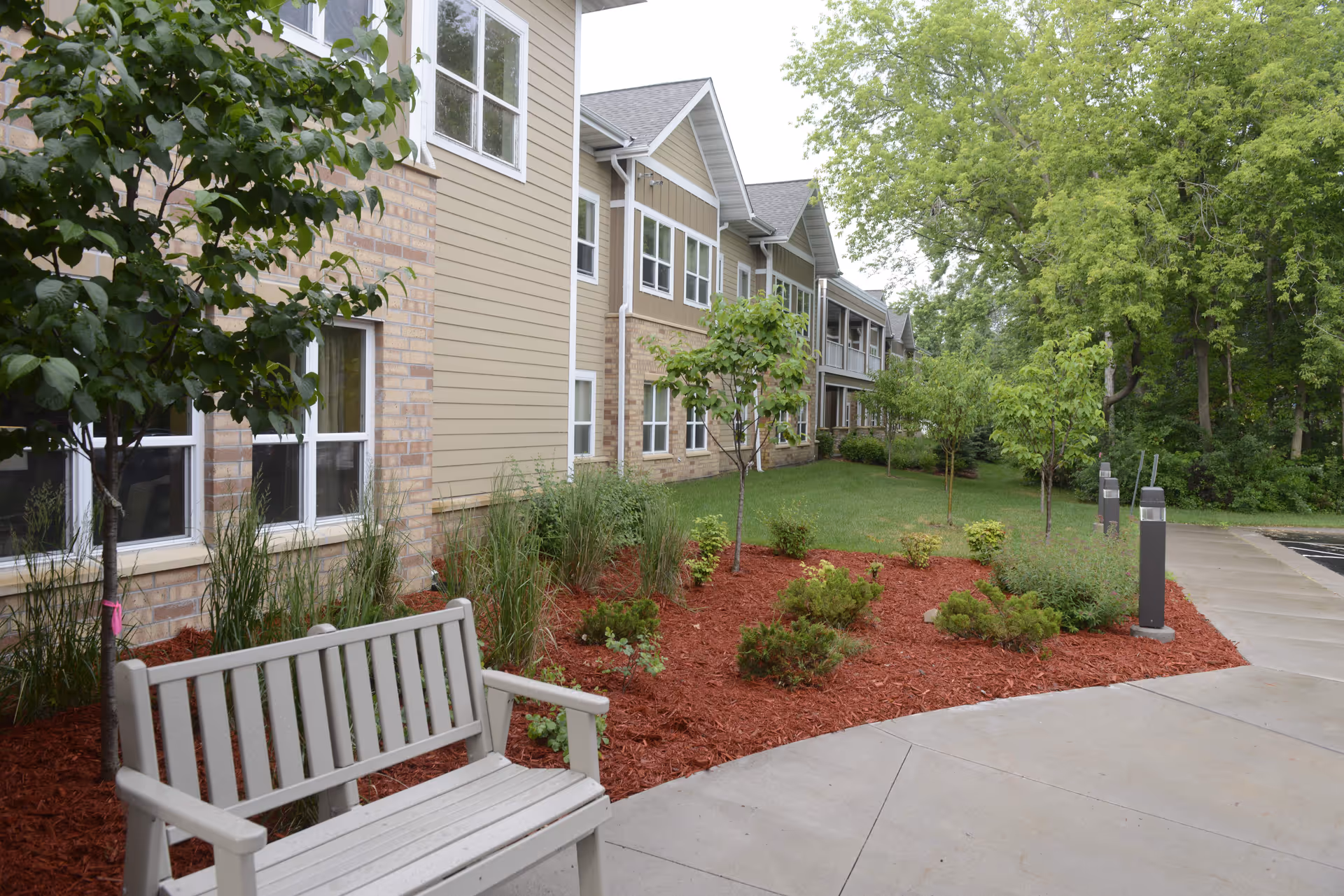 Outdoor view of a senior living facility showing a beige and brick building with multiple windows. In the foreground, there is a light gray wooden bench next to a landscaped area with small trees, bushes, and red mulch. A concrete walkway curves around the landscaped area, and there are tall trees and greenery in the background.
