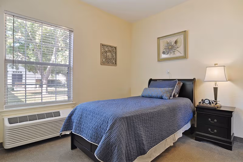 A neatly made single bed with a blue quilt and matching pillows in a senior living facility bedroom. Next to the bed is a black nightstand with a lamp, an alarm clock, and a pair of glasses. The room has beige walls with two framed artworks hanging, and a large window with white blinds letting in natural light. Below the window is a white heating and air conditioning unit.