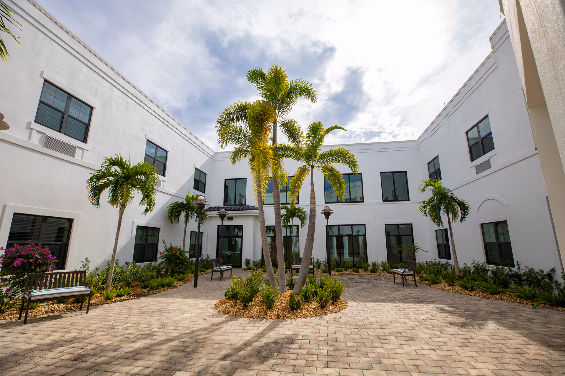 A bright courtyard area surrounded by a white two-story building with multiple windows. The courtyard features several palm trees, small plants, benches, and lamp posts under a partly cloudy sky.