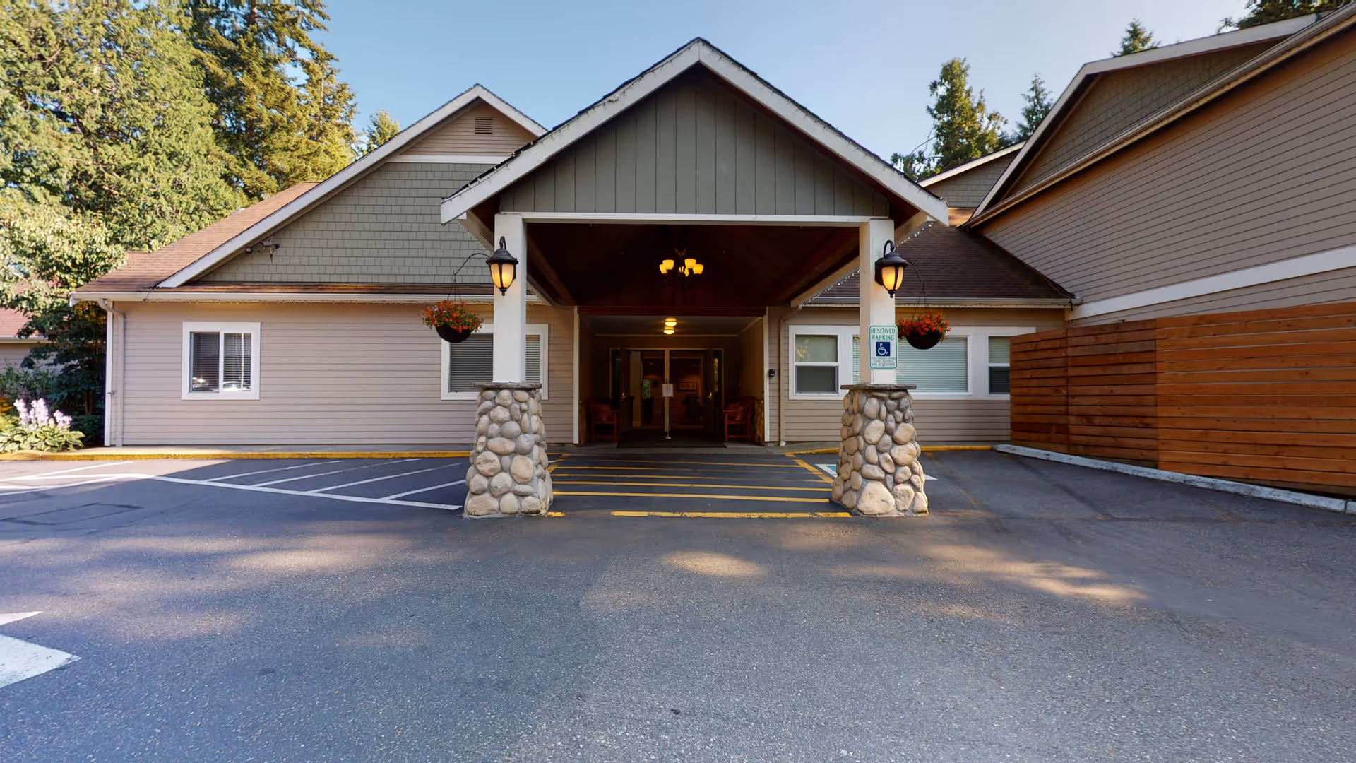 Front entrance of a single-story building with a covered driveway supported by stone pillars. The building has beige siding and a sloped roof, with hanging flower baskets on either side of the entrance. There is a designated handicapped parking space near the entrance, and trees are visible in the background.