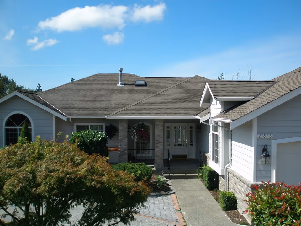 Front exterior view of a single-story residential building with a gray roof and light-colored walls. The entrance features a covered porch with two columns and a glass front door. There are bushes and trees in the landscaped front yard under a clear blue sky.