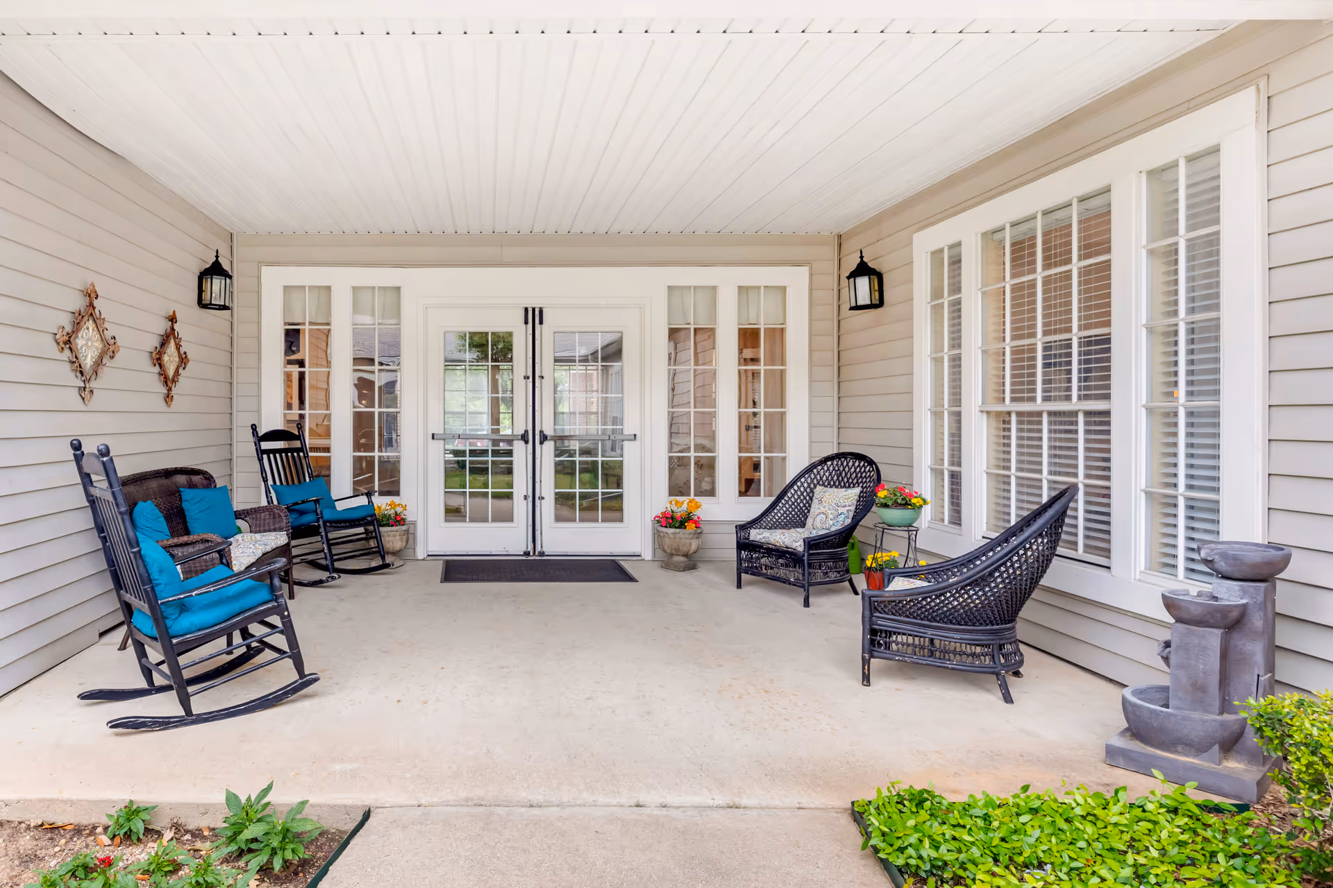 Covered outdoor patio area with two black rocking chairs with blue cushions on the left, two black wicker chairs with cushions on the right, potted flowers, and a small water fountain. The patio has beige siding walls, white ceiling, and double glass doors leading inside.
