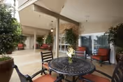 Covered outdoor patio area with a round black metal table and four chairs with red cushions. There are potted plants and greenery around the space, and additional seating with red cushions along the walls. The area is well-lit with ceiling lights and has a tiled floor.