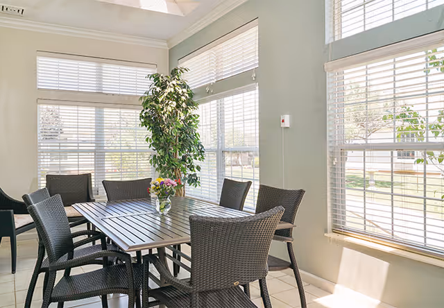 A bright dining area with a rectangular wooden table surrounded by six dark wicker chairs. Large windows with white blinds allow natural light to fill the room, and a green potted plant is placed near the corner by the windows.