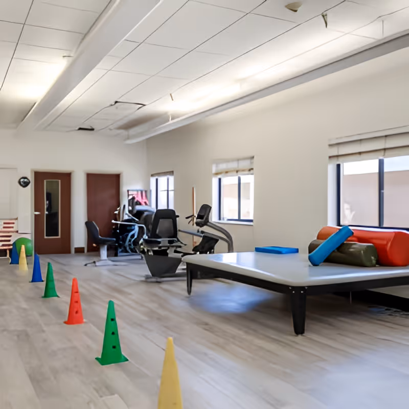 A spacious room with exercise equipment including stationary bikes and a large padded table with colorful cushions. Several colored cones are lined up on the floor, likely for physical therapy or exercise activities. The room has large windows with blinds and a white ceiling with some visible damage.