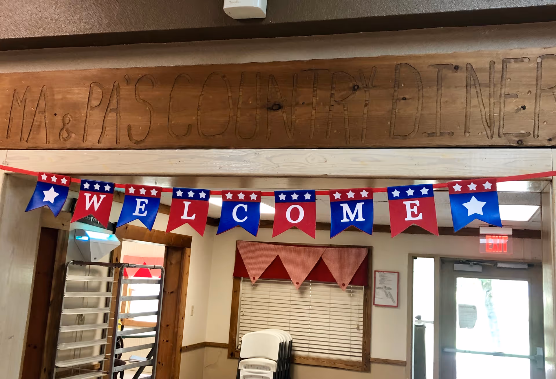 Interior view of a room with a wooden sign above the doorway that reads 'MA & PA'S COUNTRY DINER'. Below the sign, a red, white, and blue banner spells out 'WELCOME'. The room has a window with red checkered valances, stacked white chairs, and a door with an exit sign above it.