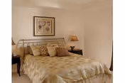 A neatly made bed with a beige and gold patterned comforter and multiple pillows in a softly lit bedroom. There are two dark wooden nightstands on either side of the bed, each with a lamp. A framed piece of artwork hangs on the wall above the bed.