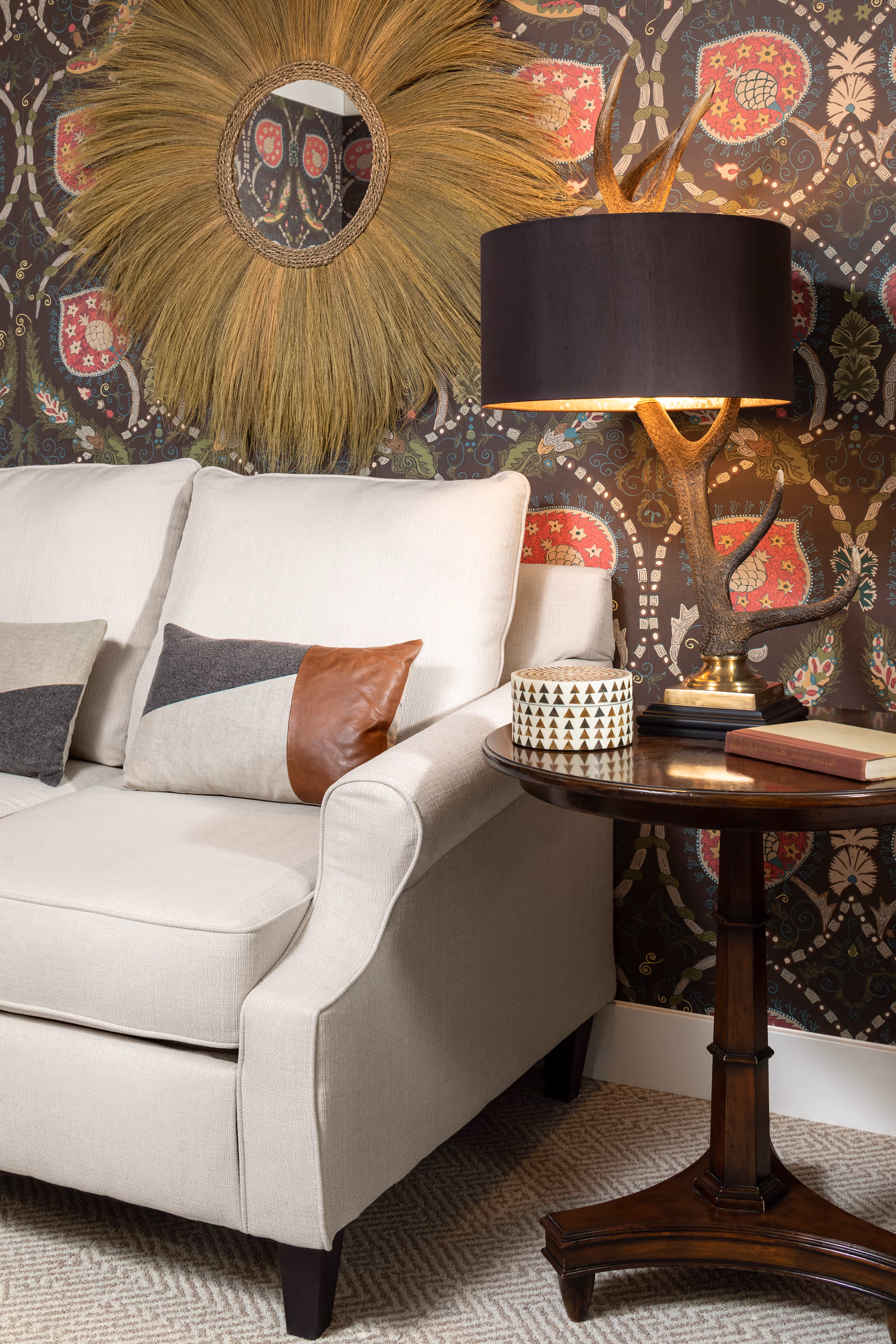 Cream sofa with geometric throw pillows next to a round wooden side table holding a lamp, books, and a decorative container in front of patterned wallpaper and a sunburst mirror.