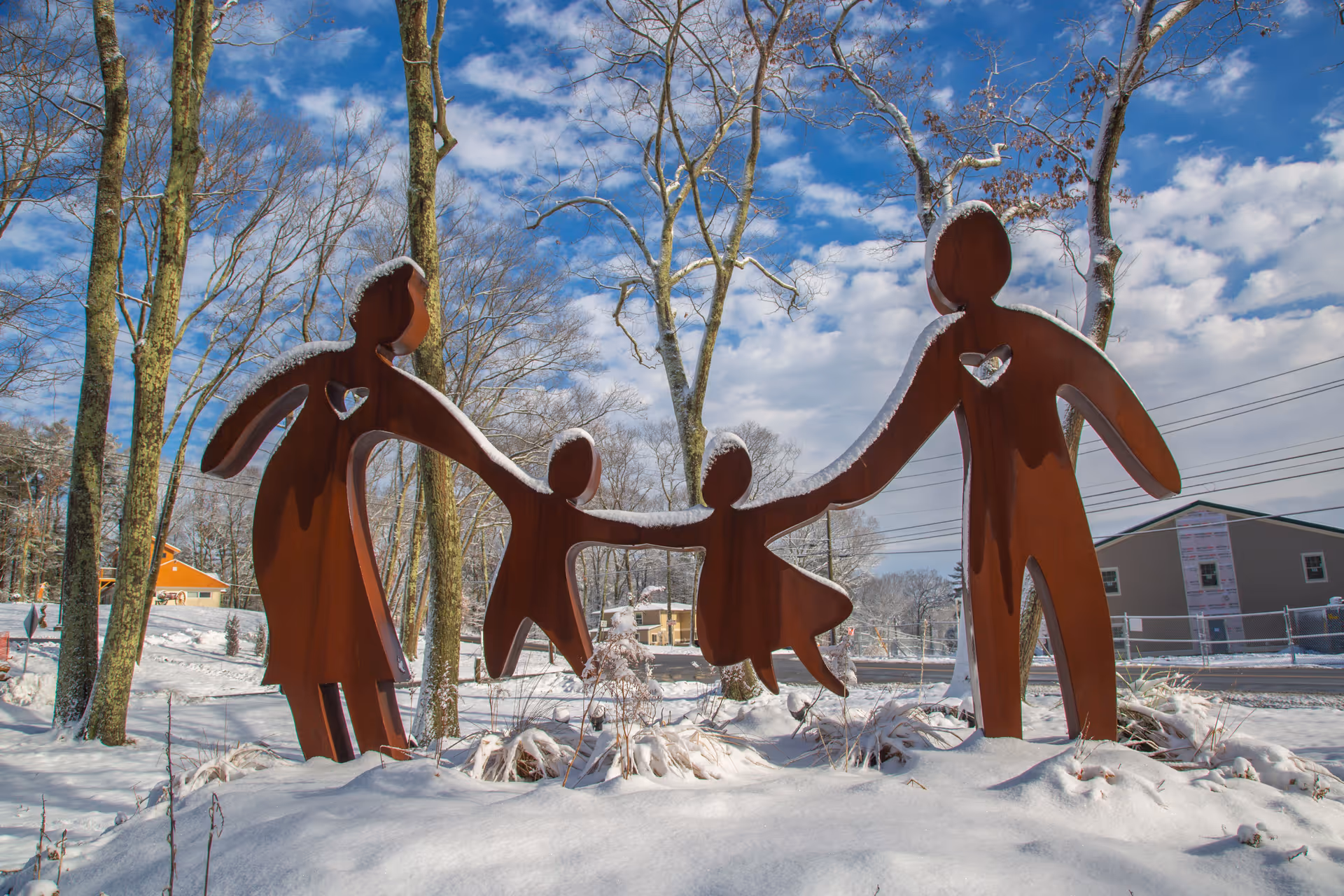 A metal sculpture of a family holding hands, consisting of two adults and two children, standing outdoors on snow-covered ground with trees and buildings in the background under a partly cloudy blue sky.
