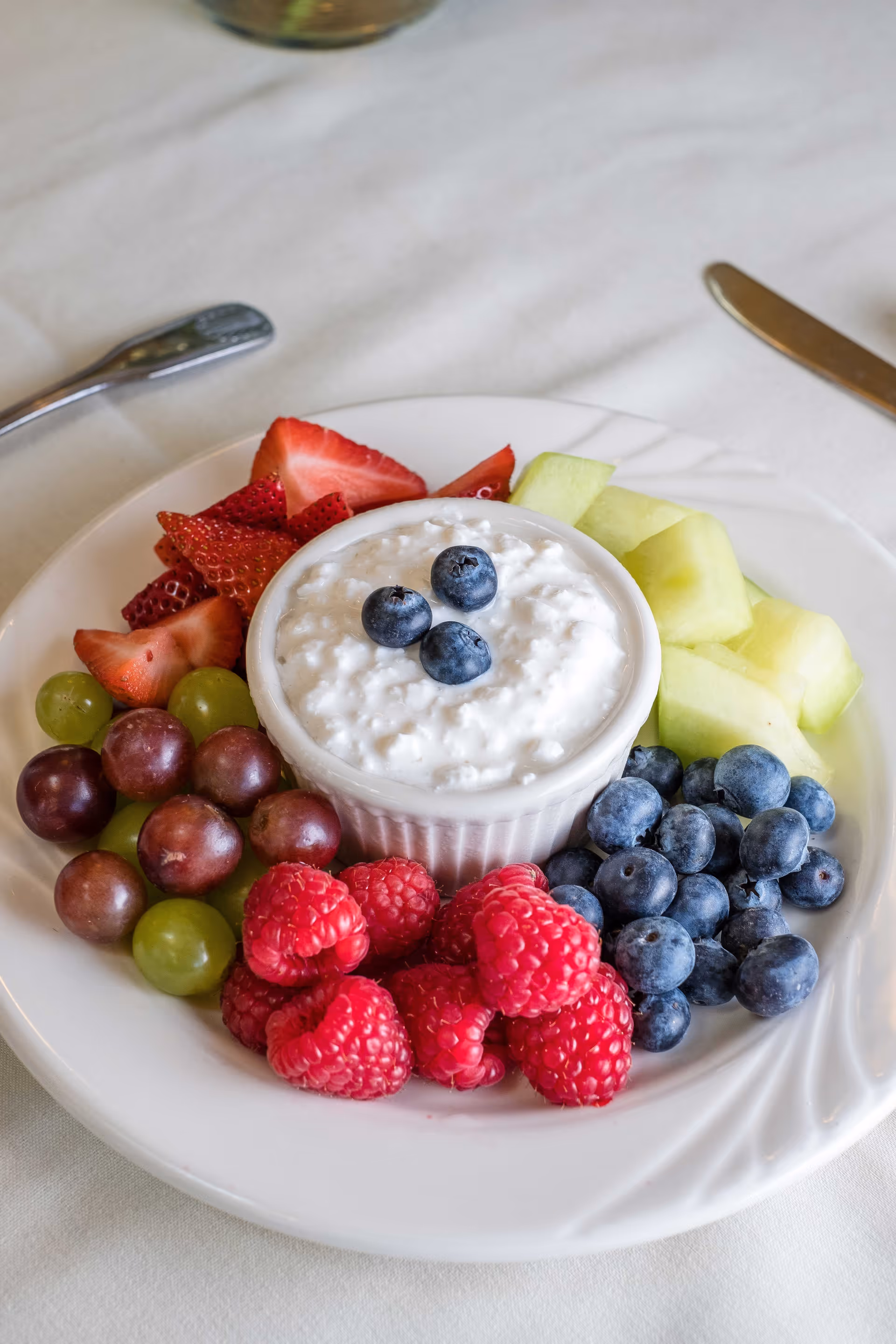 A white plate with a small white bowl of cottage cheese topped with three blueberries in the center, surrounded by fresh fruit including strawberries, green and red grapes, raspberries, blueberries, and honeydew melon cubes on a white tablecloth with a knife and spoon nearby.