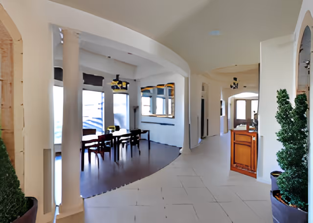 Interior view of a senior living facility showing a dining area with a table and chairs on the left, large windows with blinds, and a hallway with tiled flooring on the right. There are decorative columns and potted plants near the entrance.
