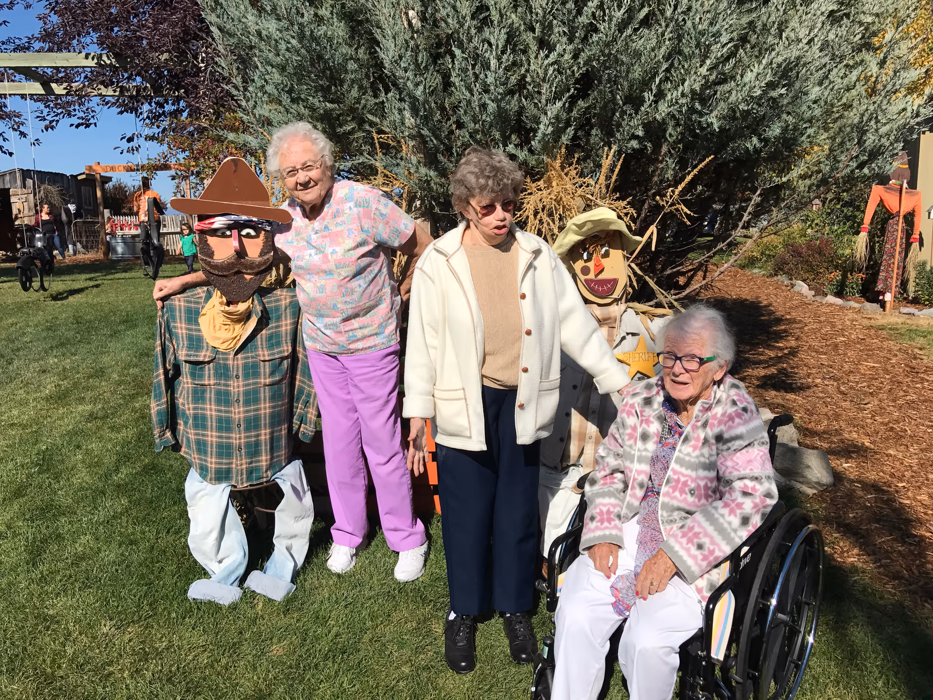 Three elderly women outdoors on a sunny day, two standing and one seated in a wheelchair, posing next to two scarecrows dressed in western attire on a grassy area with trees and garden mulch in the background.