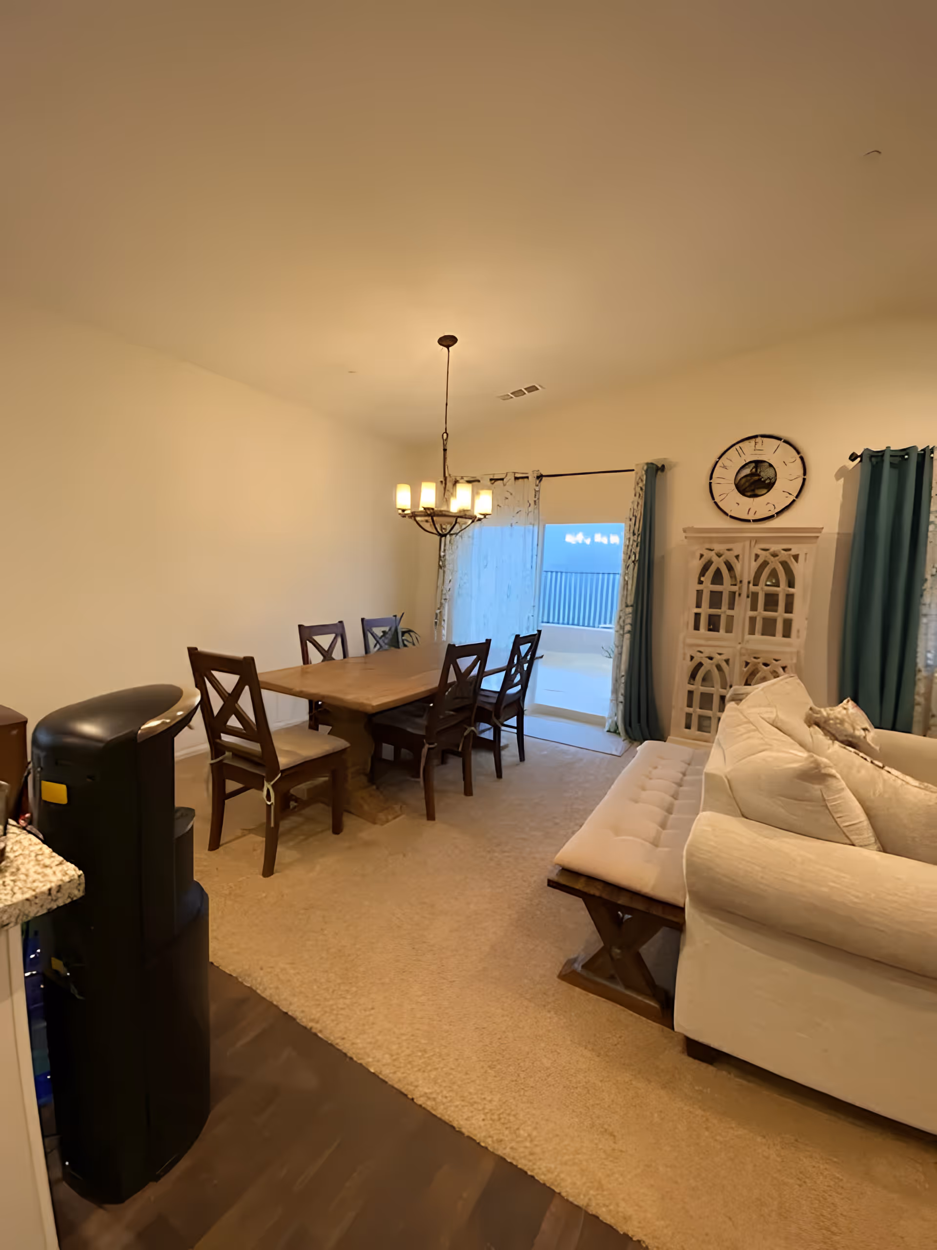 A cozy dining and living area featuring a wooden dining table with six chairs under a chandelier, a beige sofa with a cushioned bench in front, a decorative cabinet with a large clock above it, and sliding glass doors with curtains leading to an outdoor balcony.