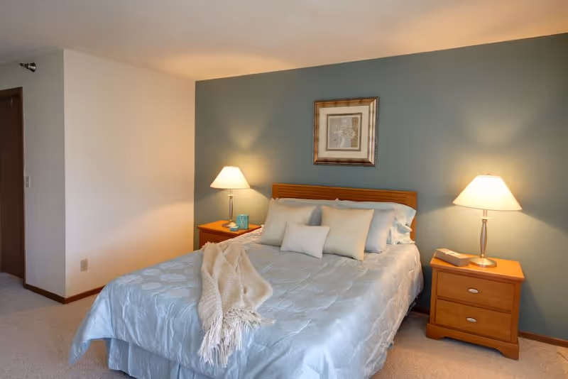 A cozy bedroom featuring a neatly made bed with light blue bedding and several pillows. There are two wooden nightstands on either side of the bed, each with a lamp. A framed artwork hangs on the teal accent wall behind the bed. The room has beige carpeting and light-colored walls.