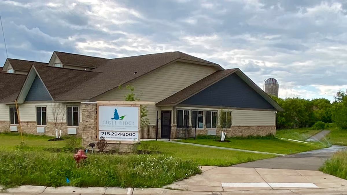 Exterior view of Eagle Ridge Senior Living facility showing a single-story building with beige and blue siding, a stone base, and a brown roof. There is a sign in front with the facility name, logo, and phone number. The surrounding area includes green grass, a sidewalk, and a cloudy sky.