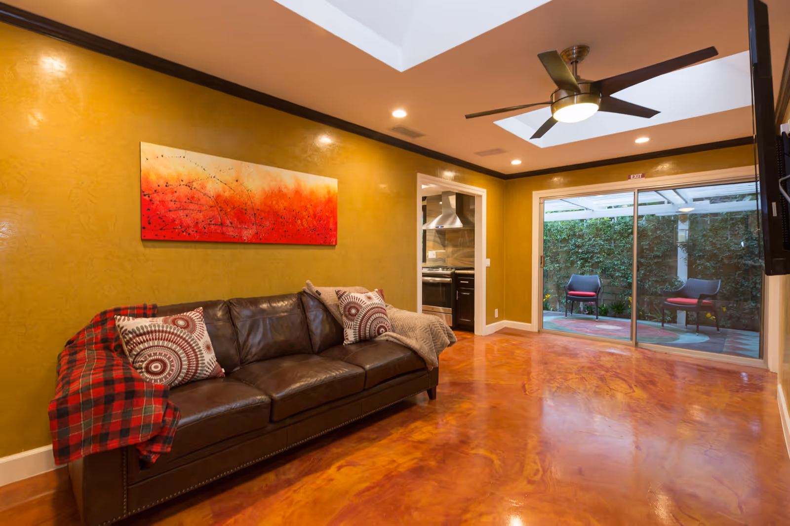A cozy living room with a dark brown leather sofa adorned with patterned pillows and a red plaid blanket. The walls are painted mustard yellow with a large abstract painting featuring red and orange hues. The room has a polished reddish-brown floor, a ceiling fan with lights, recessed lighting, and a skylight. A sliding glass door leads to an outdoor patio with two chairs and greenery.