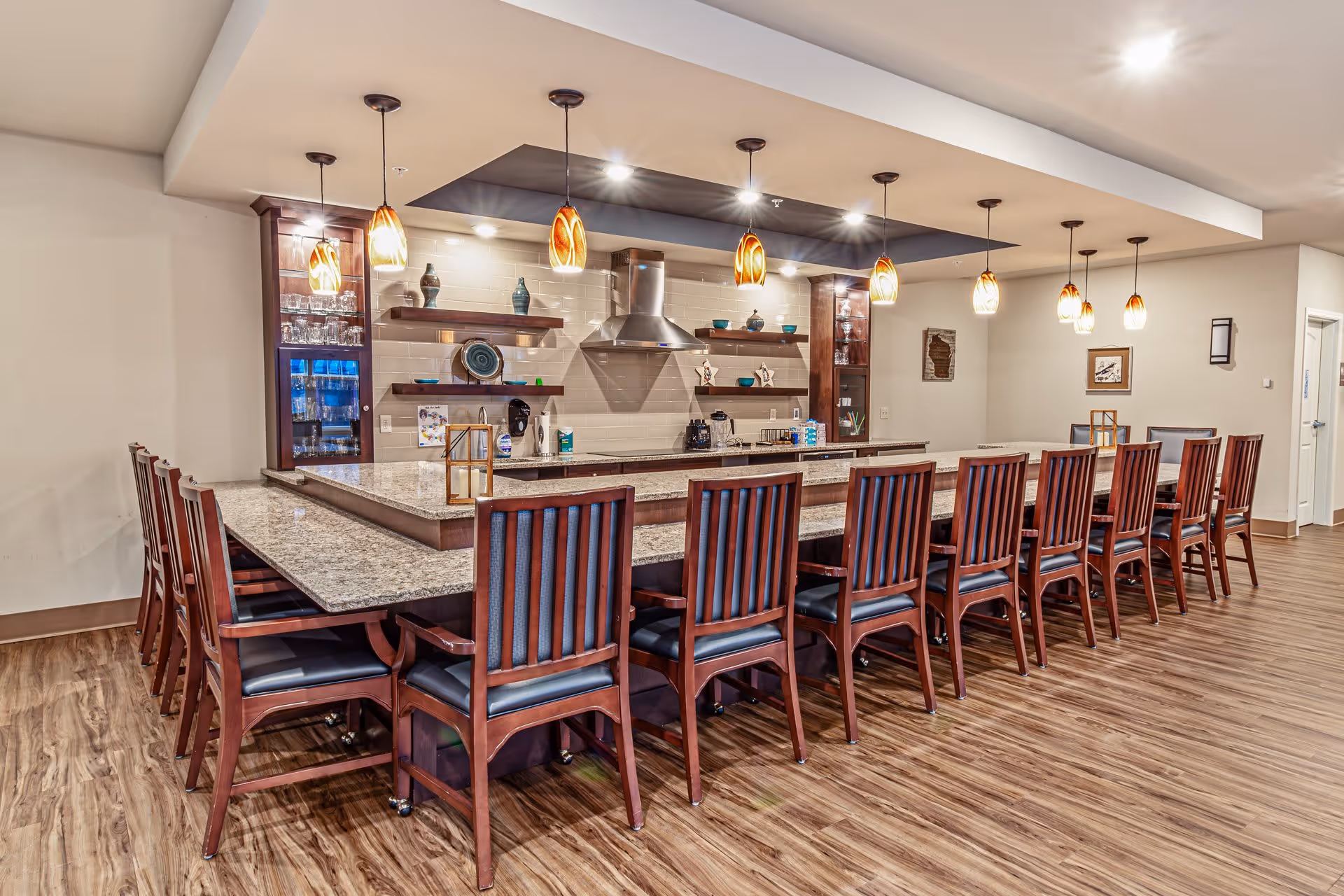 A large kitchen area with a long granite countertop surrounded by wooden chairs with black cushions. The kitchen features modern pendant lights hanging from the ceiling, open shelves with decorative items, a stainless steel range hood, and wooden cabinetry. The floor is wood-patterned, and the space is well-lit with recessed lighting.