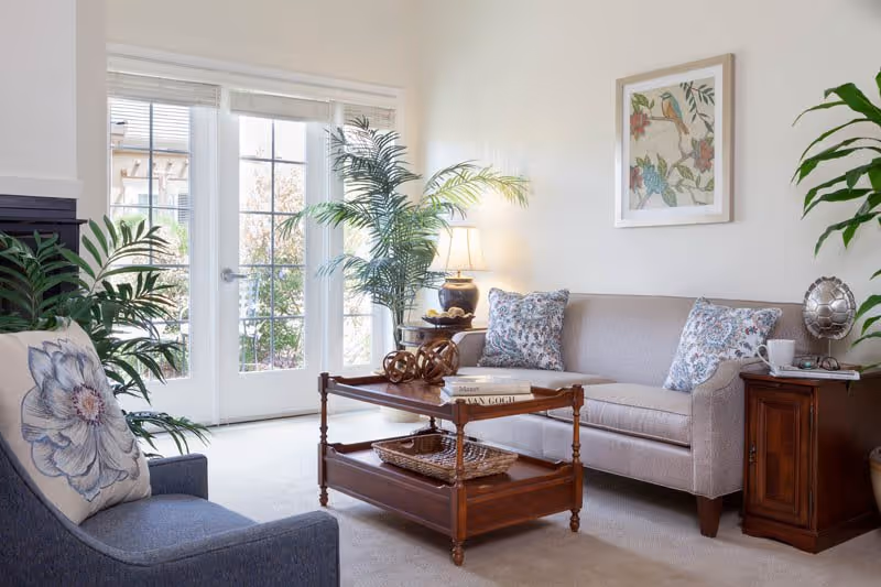 A bright living room with a beige sofa adorned with floral patterned pillows, a wooden coffee table with books and decorative items, a blue armchair with a floral cushion, potted plants, a table lamp, and a framed bird artwork on the wall. French doors with glass panes open to an outdoor view.