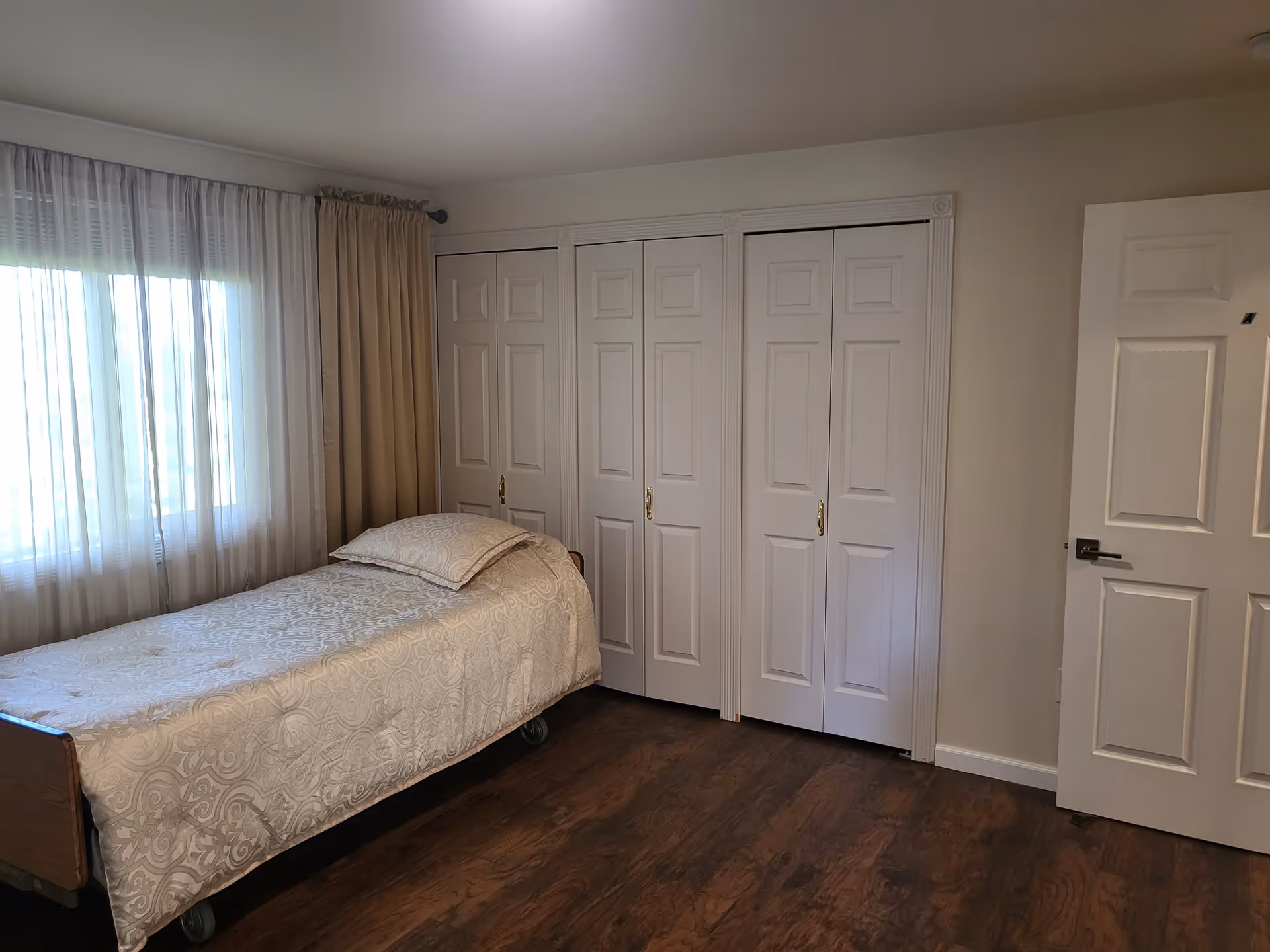 A simple bedroom with a single bed covered in a beige patterned bedspread and pillow. The room has wooden flooring, a window with sheer and beige curtains, and a set of white closet doors along one wall. There is also a white door partially open on the right side of the image.