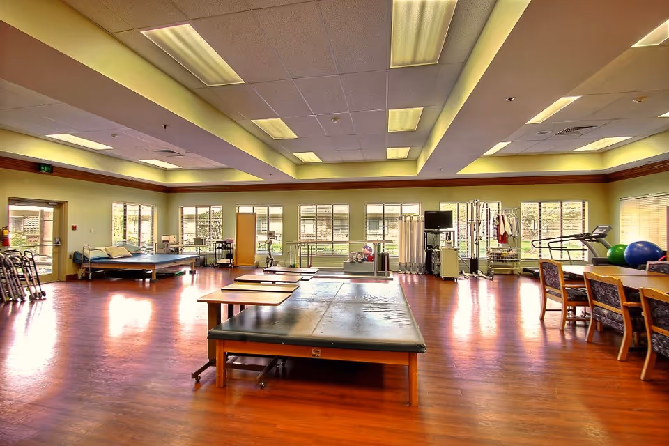 A spacious rehabilitation or physical therapy room with wooden floors, padded therapy tables, exercise equipment including a treadmill and therapy balls, chairs, and large windows allowing natural light to fill the room.