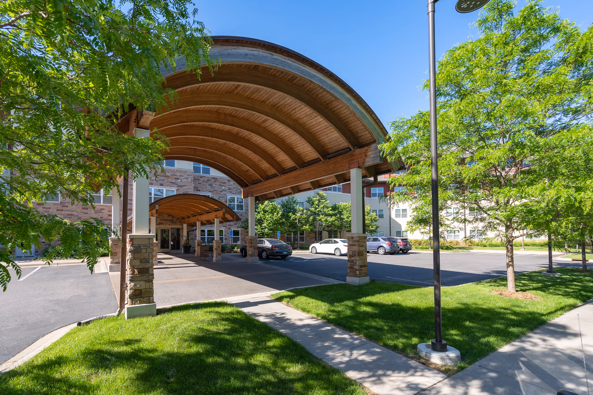 Entrance area of The Glenn Minnetonka facility featuring a covered driveway with wooden arched roof supported by stone pillars, surrounded by green trees and a parking lot with several parked cars under a clear blue sky.
