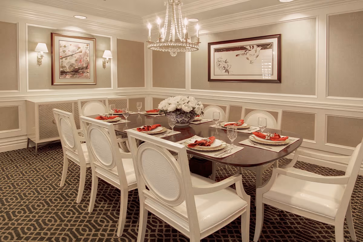 Elegant dining room with a dark wooden table set for eight people. Each place setting includes a white plate, a red napkin, silverware, and a clear glass. The room features white paneled walls with beige fabric inserts, two framed artworks, wall sconces, a patterned carpet, and a crystal chandelier hanging above the table. A floral centerpiece is placed in the middle of the table.