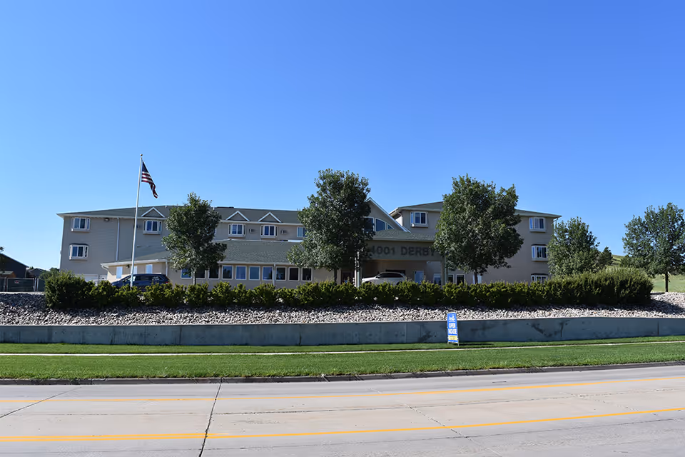 Exterior view of a multi-story senior living facility building with beige siding and green roofing. There are several trees and bushes in front of the building, an American flag on a flagpole, and a blue open house sign near the road. The sky is clear and blue.