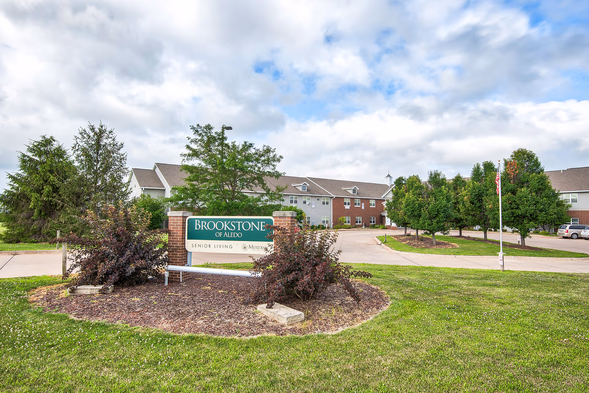Exterior view of Brookstone of Aledo senior living facility with a large green and white sign in front surrounded by bushes and trees. The building is two stories with a mix of brick and siding, and there is a driveway and parking area visible. The sky is partly cloudy.