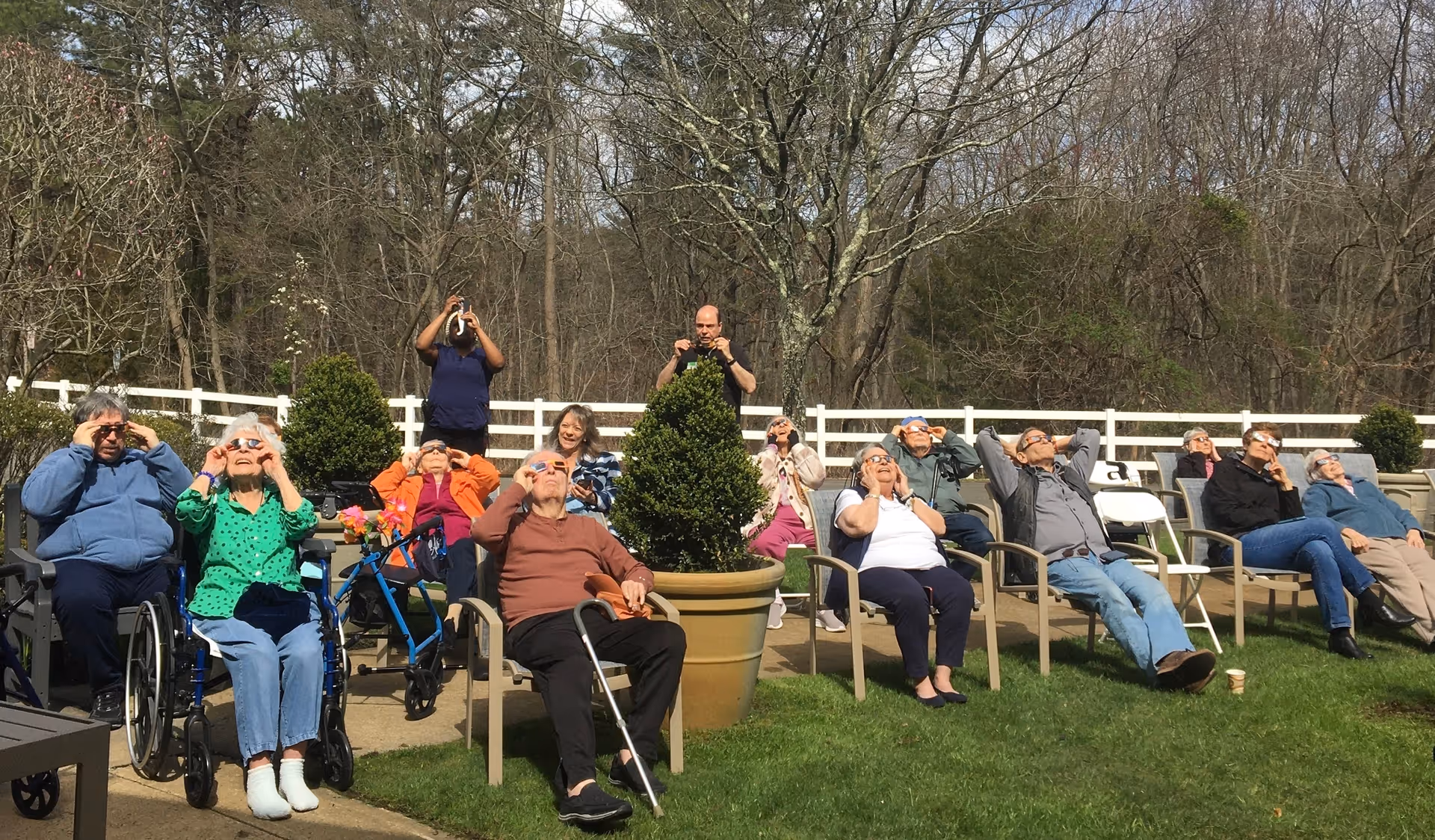 A group of elderly people sitting outdoors on chairs and wheelchairs in a garden area, all looking up at the sky wearing solar eclipse glasses. Two people are standing behind them taking photos. The scene is sunny with trees and a white fence in the background.