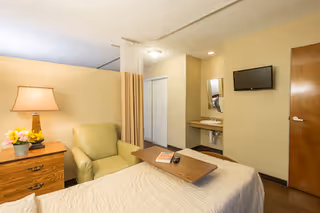 A nursing facility room featuring a bed with a wooden tray holding a remote and a book, a green armchair, a wooden nightstand with a lamp and flowers, a wall-mounted TV, a small sink with a mirror, and a closed wooden door.