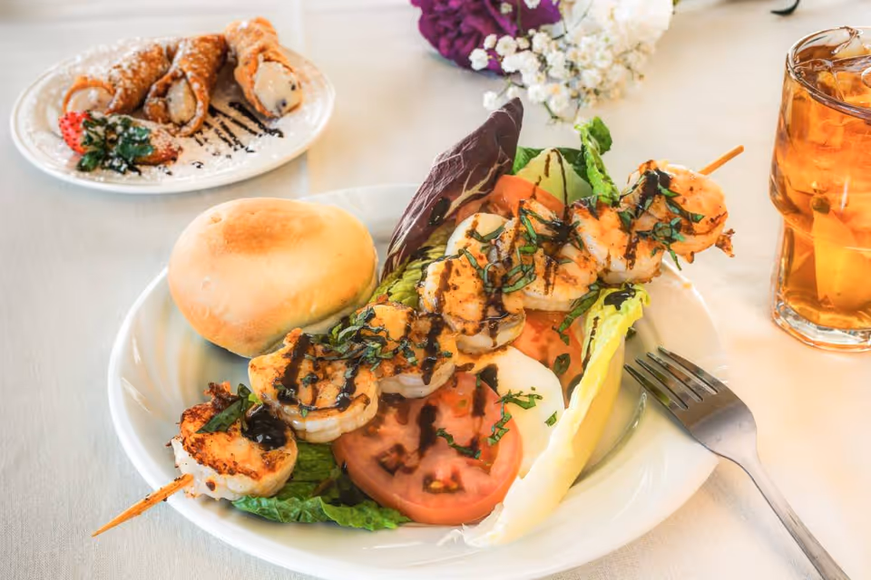 A plate with grilled shrimp skewers served on a bed of lettuce and tomato slices, accompanied by a bread roll. In the background, there is a plate with three cannoli and a strawberry, and a glass of iced tea with lemon on the right side. A fork is placed next to the plate with shrimp.