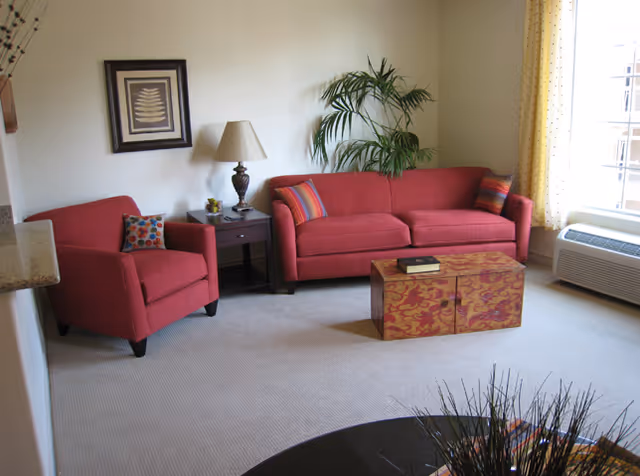 A cozy living room with a red sofa and matching armchair, both adorned with colorful throw pillows. A wooden coffee table with a decorative pattern sits in front of the sofa, with a book on top. A side table with a lamp and small decorative items is placed between the sofa and armchair. A large green plant is positioned in the corner near a window with yellow curtains, and a framed artwork hangs on the wall above the armchair. The room has beige carpeting and an air conditioning unit below the window.