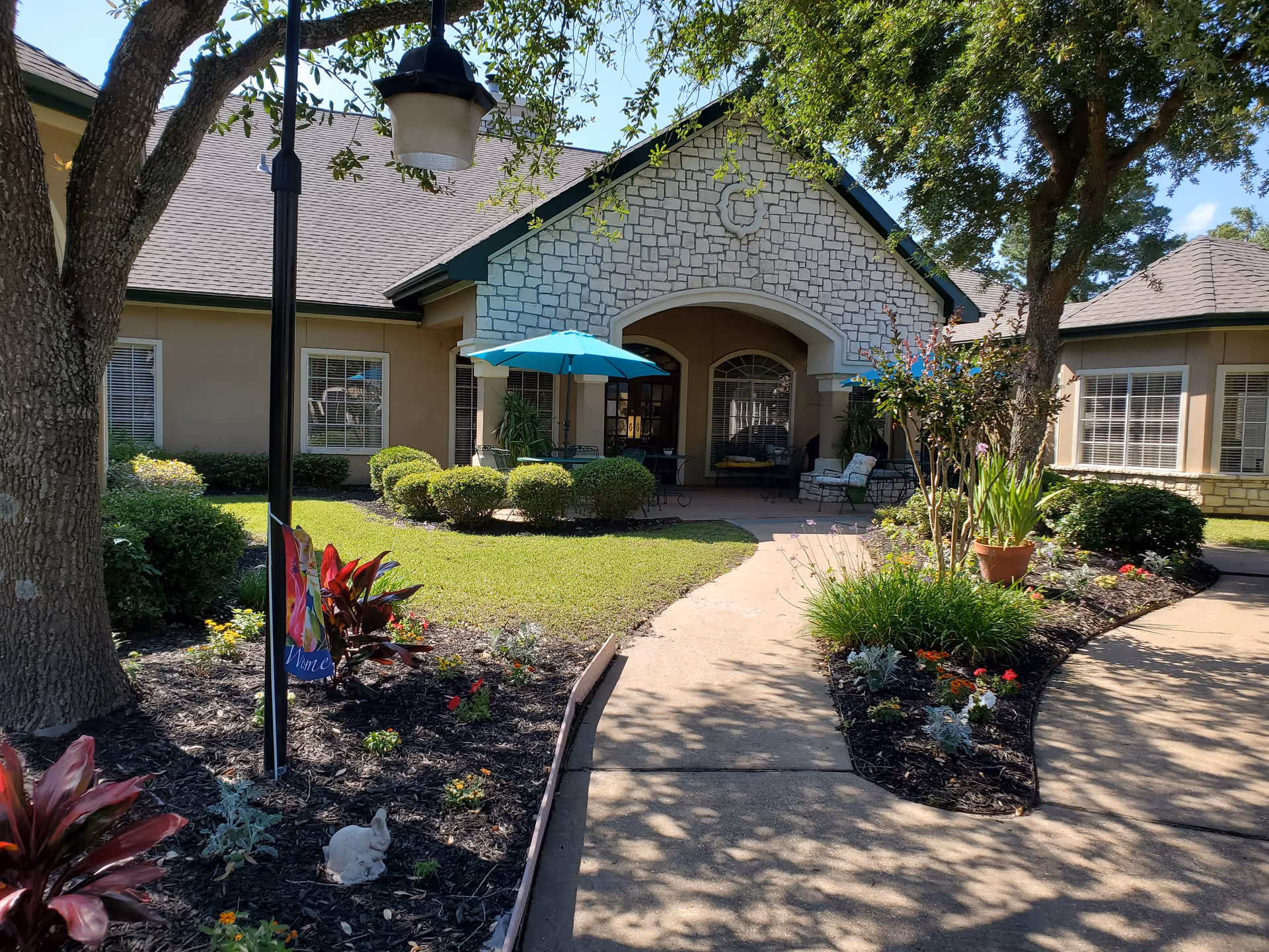Outdoor view of Carriage Inn Huntsville featuring a paved walkway leading to a building entrance with stone and beige walls. The area is landscaped with green bushes, colorful flowers, and trees providing shade. There are patio tables with blue umbrellas and outdoor seating near the entrance.