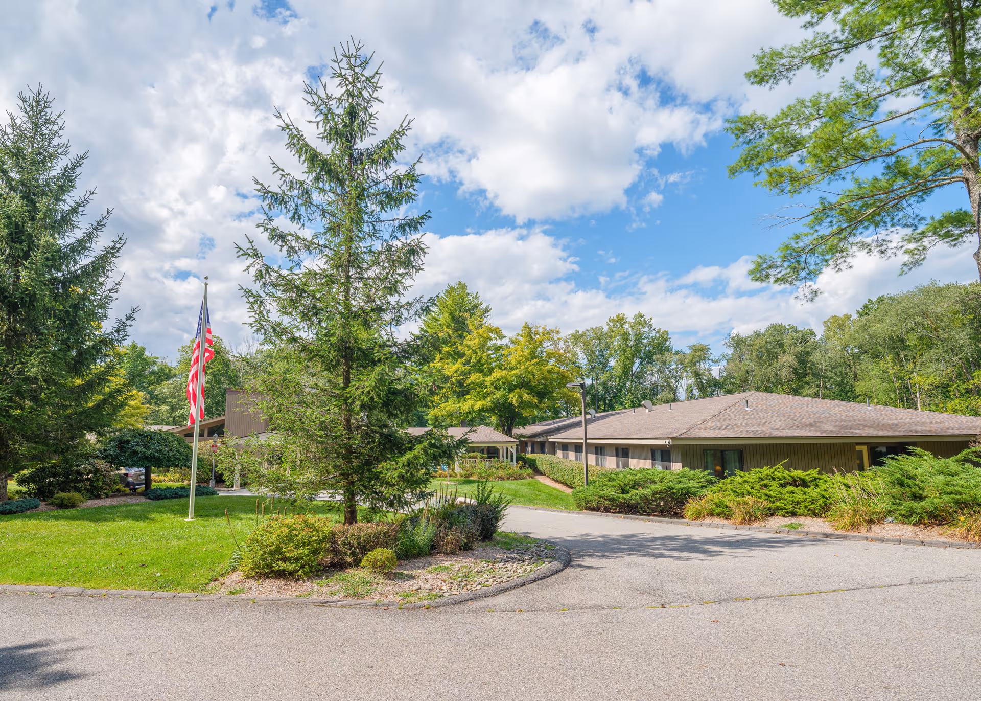 Exterior view of The Country House in Westchester facility showing a single-story building surrounded by green trees and shrubs under a partly cloudy sky. An American flag is displayed on a flagpole near the driveway entrance.