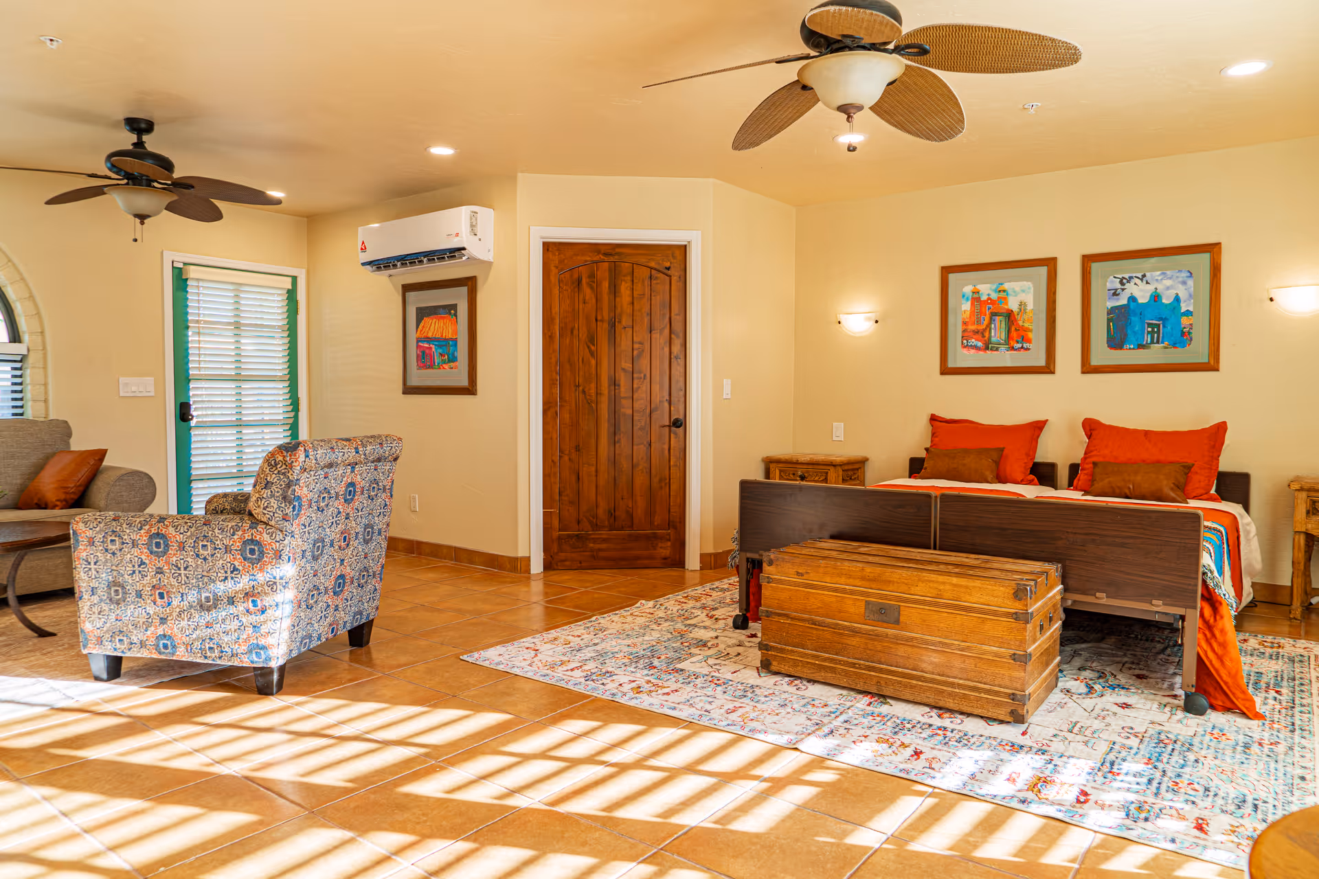 A bright and spacious bedroom in an assisted living facility featuring a bed with red and brown pillows, a wooden chest at the foot of the bed, colorful framed artwork on the wall, a patterned armchair, a sofa, ceiling fans, and a door with a green frame leading outside. The room has tiled floors and a large area rug under the bed.