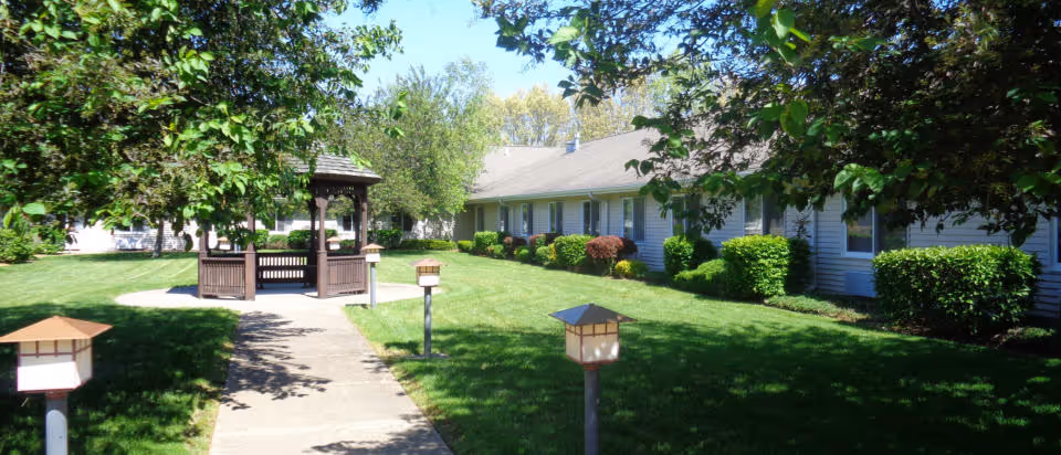 A well-maintained outdoor garden area at Liberty Village of Streator featuring a concrete walkway leading to a wooden gazebo surrounded by green grass, bushes, and trees under a clear sky.