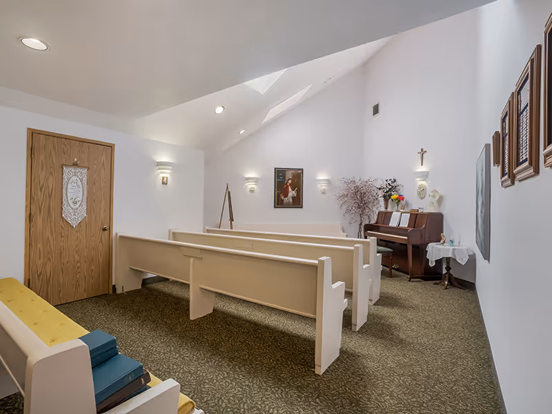 Interior view of a small chapel or prayer room with white walls and a slanted ceiling with skylights. The room contains several white wooden pews arranged in rows, a wooden piano with sheet music, a small table with a white cloth, flowers, and religious items. There are framed pictures and a cross on the walls, and a wooden door on the left side.