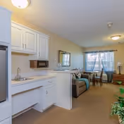 Interior view of a senior living facility apartment at Alpine House showing a small kitchen area with white cabinets, a sink, and a microwave. The living area includes a sofa with cushions, a dining table with chairs near a window with blue curtains, and a wooden dresser with a lamp. The room is well-lit with ceiling lights and has beige carpeting.