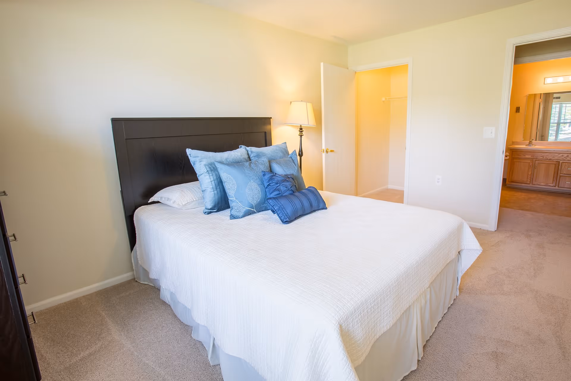 A bedroom with a neatly made bed featuring a white bedspread and several blue decorative pillows. There is a dark wooden headboard and a floor lamp beside the bed. The room has beige carpet and light-colored walls. Two open doorways are visible, one leading to a walk-in closet and the other to a bathroom with a wooden vanity and mirror.