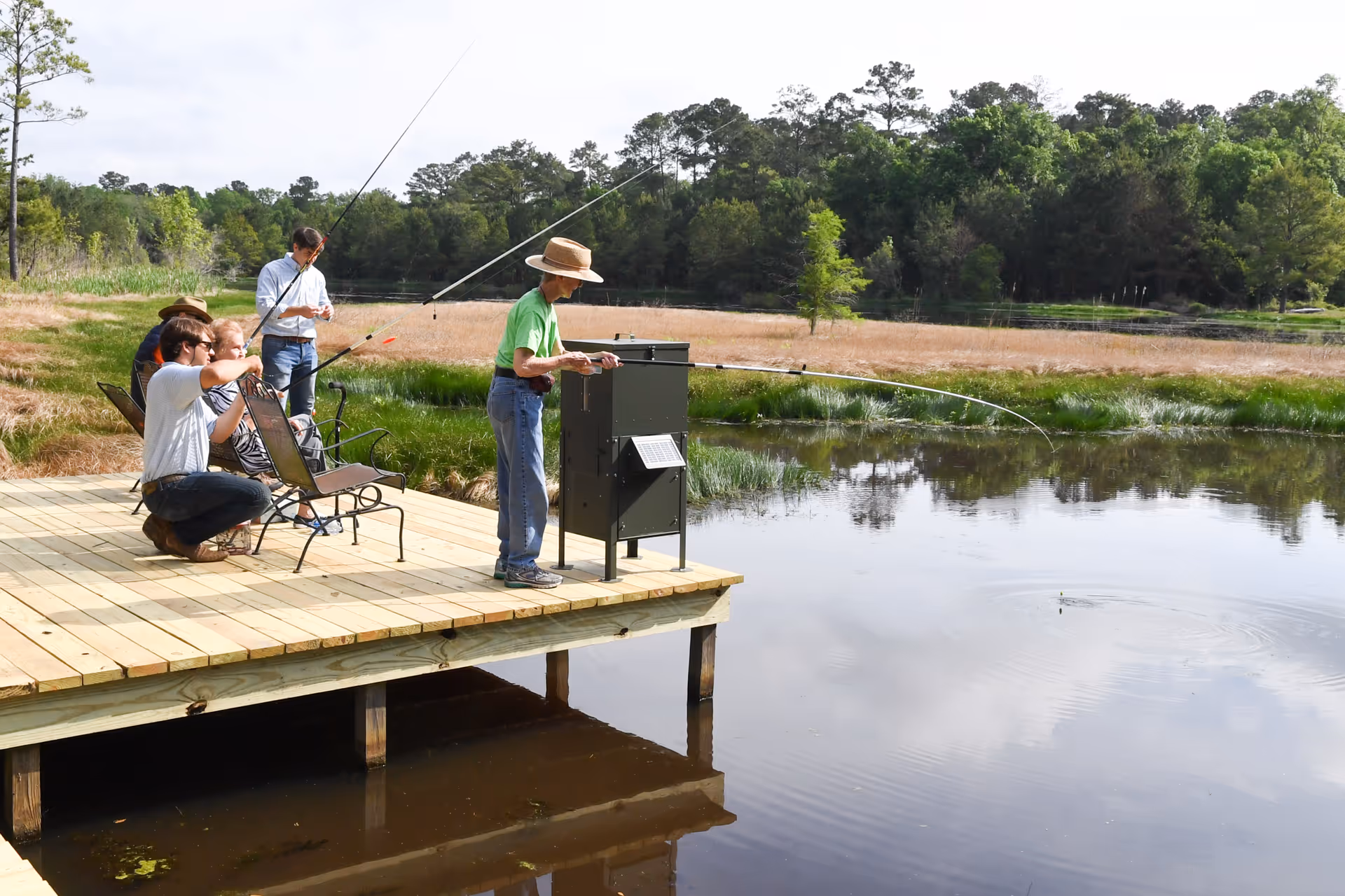 A group of people fishing on a wooden dock by a calm pond surrounded by trees and grass under a partly cloudy sky.