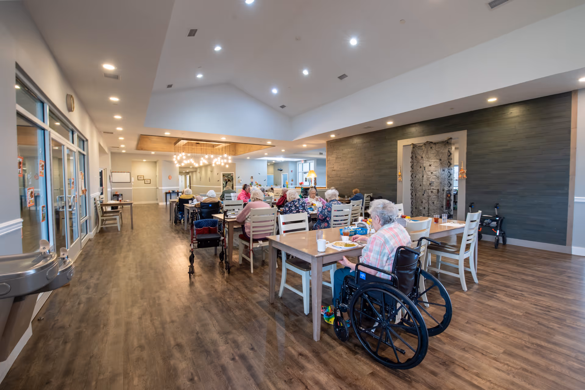 A communal dining area in a senior living facility with residents seated at tables, some in wheelchairs, wood floors, and overhead lighting.