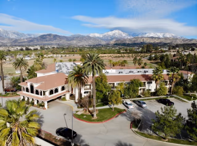 Aerial view of The Village at Redlands senior living facility showing a large building with a red-tiled roof surrounded by palm trees and greenery. There is a parking lot with several cars in front of the building. In the background, there are open fields and snow-capped mountains under a partly cloudy sky.