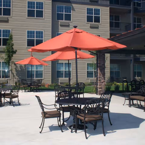 Outdoor patio area with several black metal tables and chairs, each table shaded by large red umbrellas. The patio is adjacent to a multi-story building with multiple windows and some greenery around the edges.
