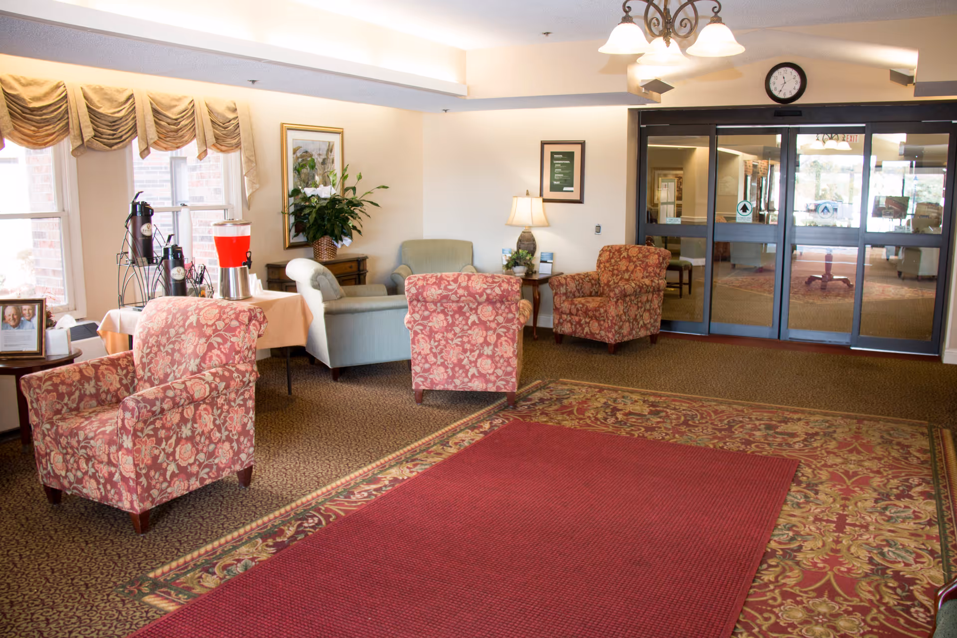 A senior living facility lobby with floral armchairs and sofas arranged around side tables, a beverage station by the windows, and glass automatic entrance doors.