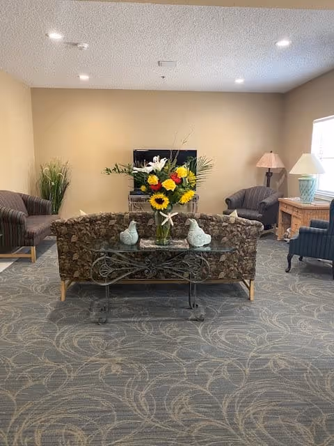 A cozy living room area with a floral patterned sofa facing a TV on a stand. There is a glass table behind the sofa with a vase of colorful flowers and two decorative bird figurines. The room has a patterned carpet, beige walls, and several armchairs. Two table lamps are placed on wooden side tables near the windows.